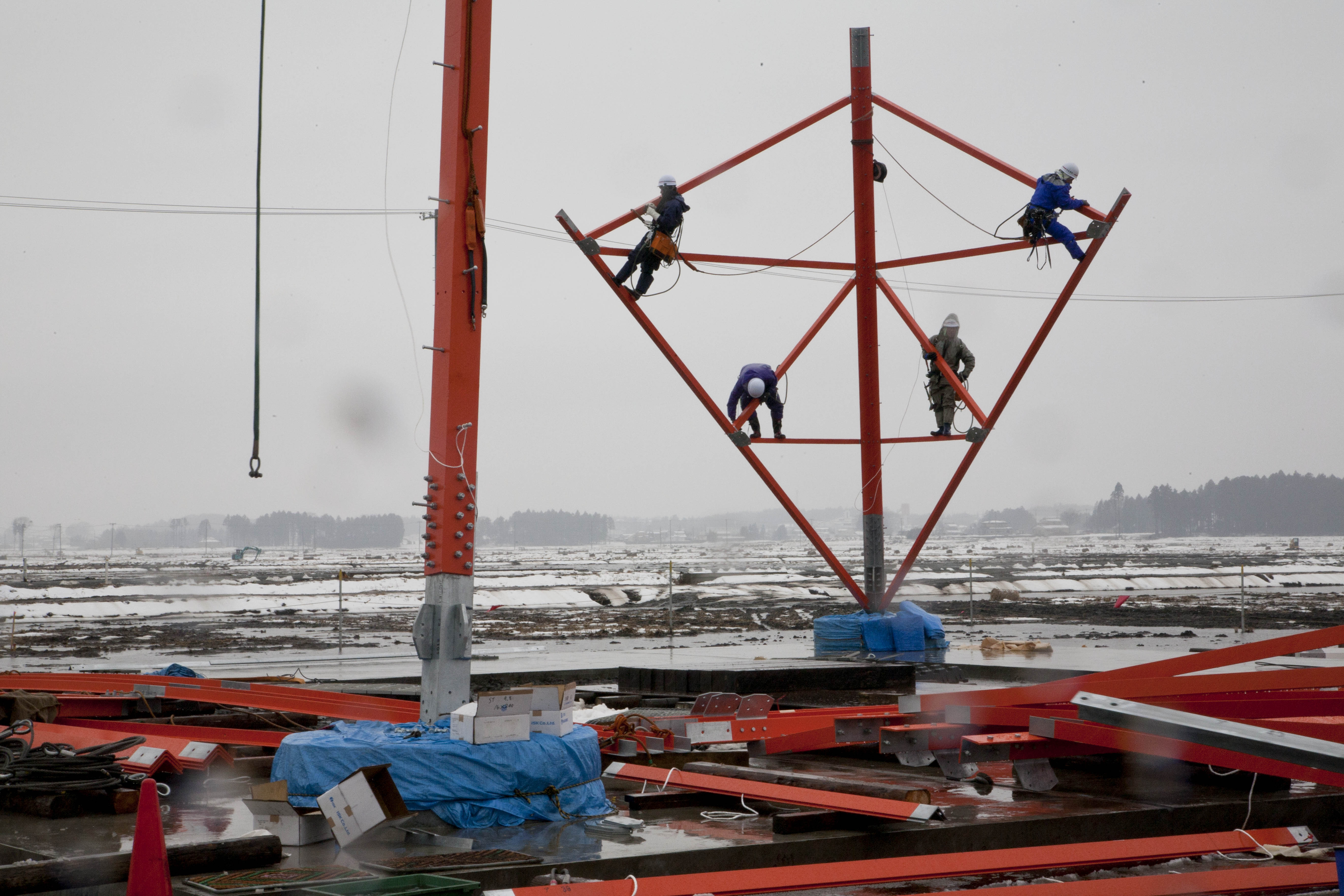 Par gros temps, les ouvriers assemblent un pylone pour le transport de l'électricité à haute-tension. Il y a un an, le tsunami détruisait les habitations et les rizières à cet endroit et applatissait le paysage.